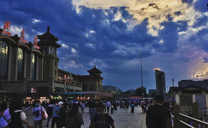 Beijing Railway Station under a bright, orange sun scattered by billowing, blue clouds.